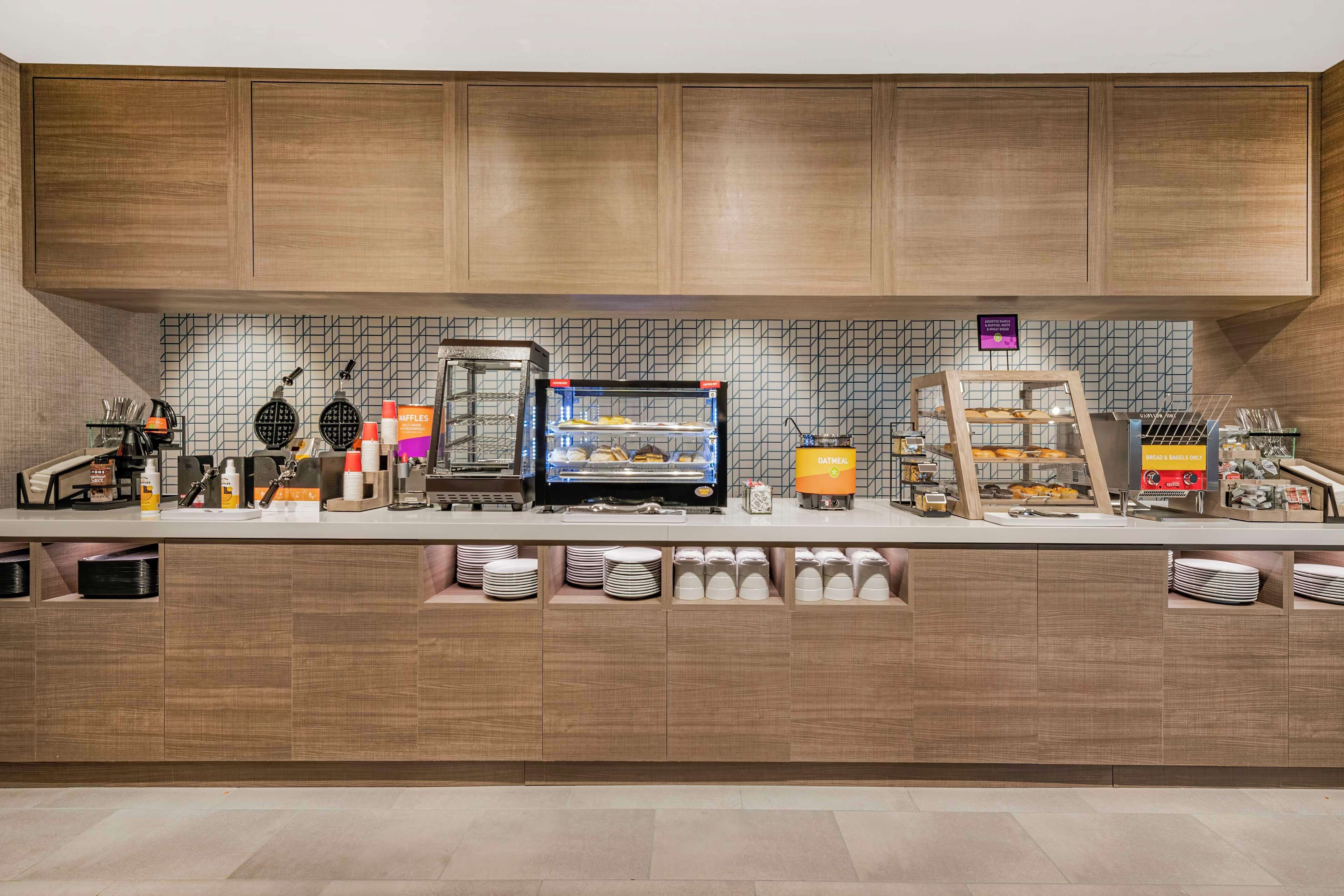 A breakfast bar with a waffle maker, coffee machine, and a display case with pastries.