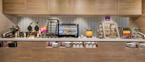 A breakfast bar with a waffle maker, coffee machine, and a display case with pastries.