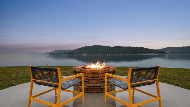 A fire pit with two chairs facing it, overlooking a calm lake with distant hills. - Home2 Suites By Hilton Guntersville (Guntersville)
