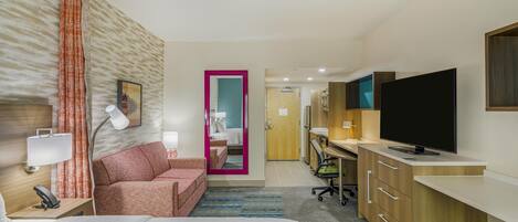 Habitación de hotel con cama, escritorio, televisor, lámpara de escritorio, silla, sofá, baño con espejo y puerta.