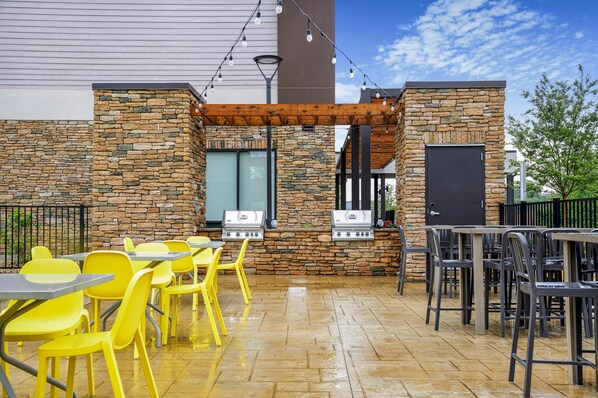 An outdoor patio with stone walls, grilling stations, yellow chairs, and tables. - Home2 Suites By Hilton Guntersville (Guntersville)