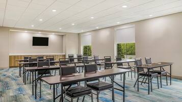 A conference room with multiple tables and chairs, a flat-screen TV mounted on the wall, and large windows with a view of greenery.