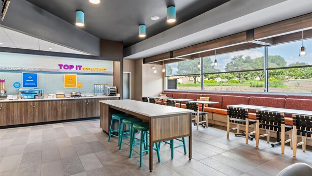 A modern cafe with a long counter, seating area, and large windows offering a view of the outdoors. - Tru By Hilton Kansas City Airport (Kansas City)