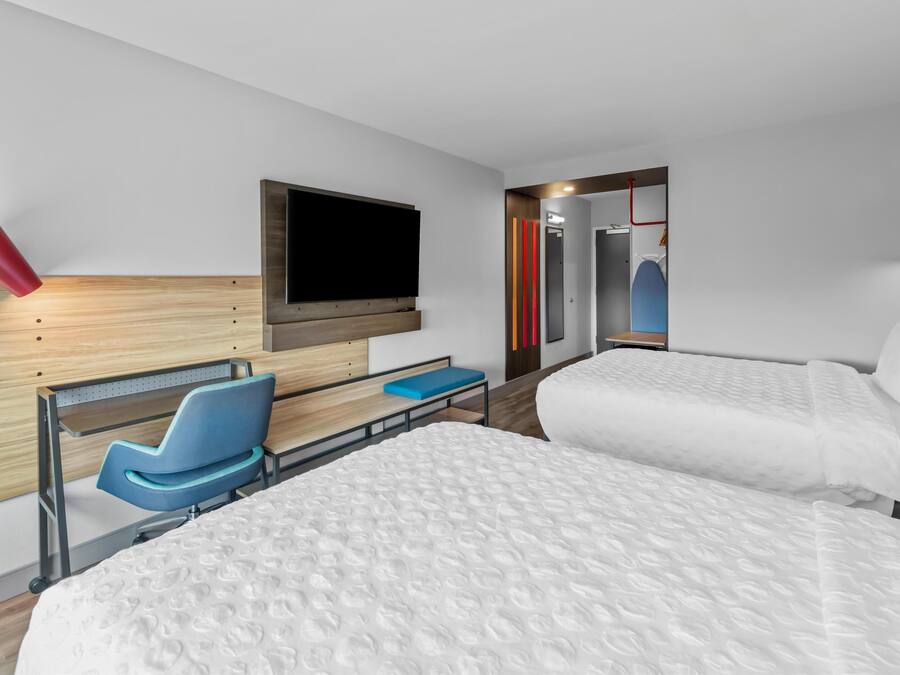 Habitación de hotel con dos camas, un televisor montado en la pared, una silla azul y un cabecero de madera.