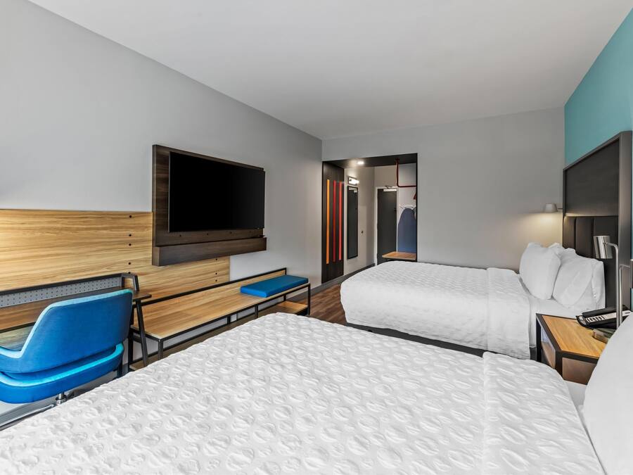 A hotel room with two beds, a TV mounted on the wall, a desk with a chair, and a small table with a keyboard.