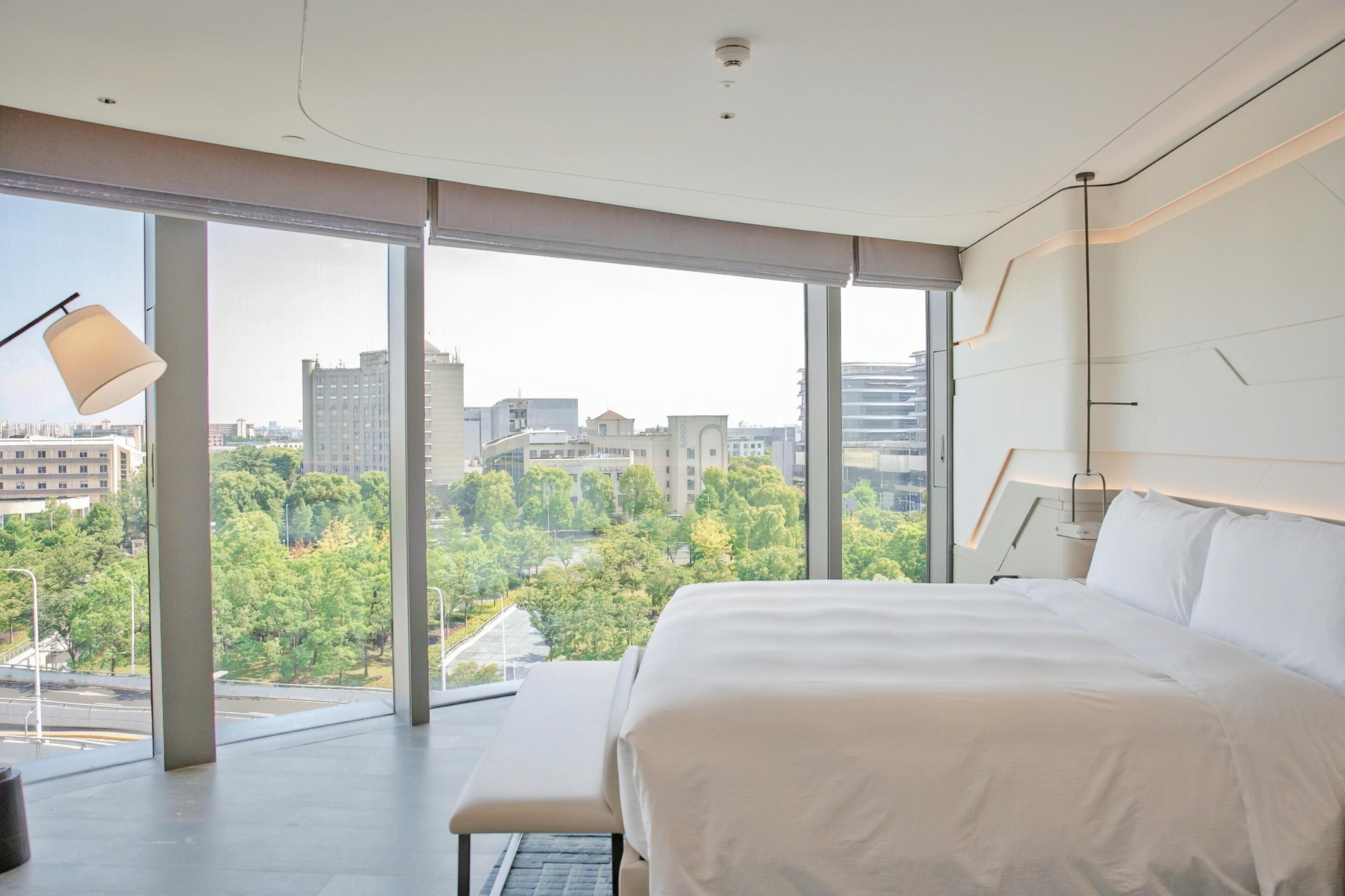 A modern hotel room with a large bed, a bench, and a view of the cityscape through floor-to-ceiling windows.