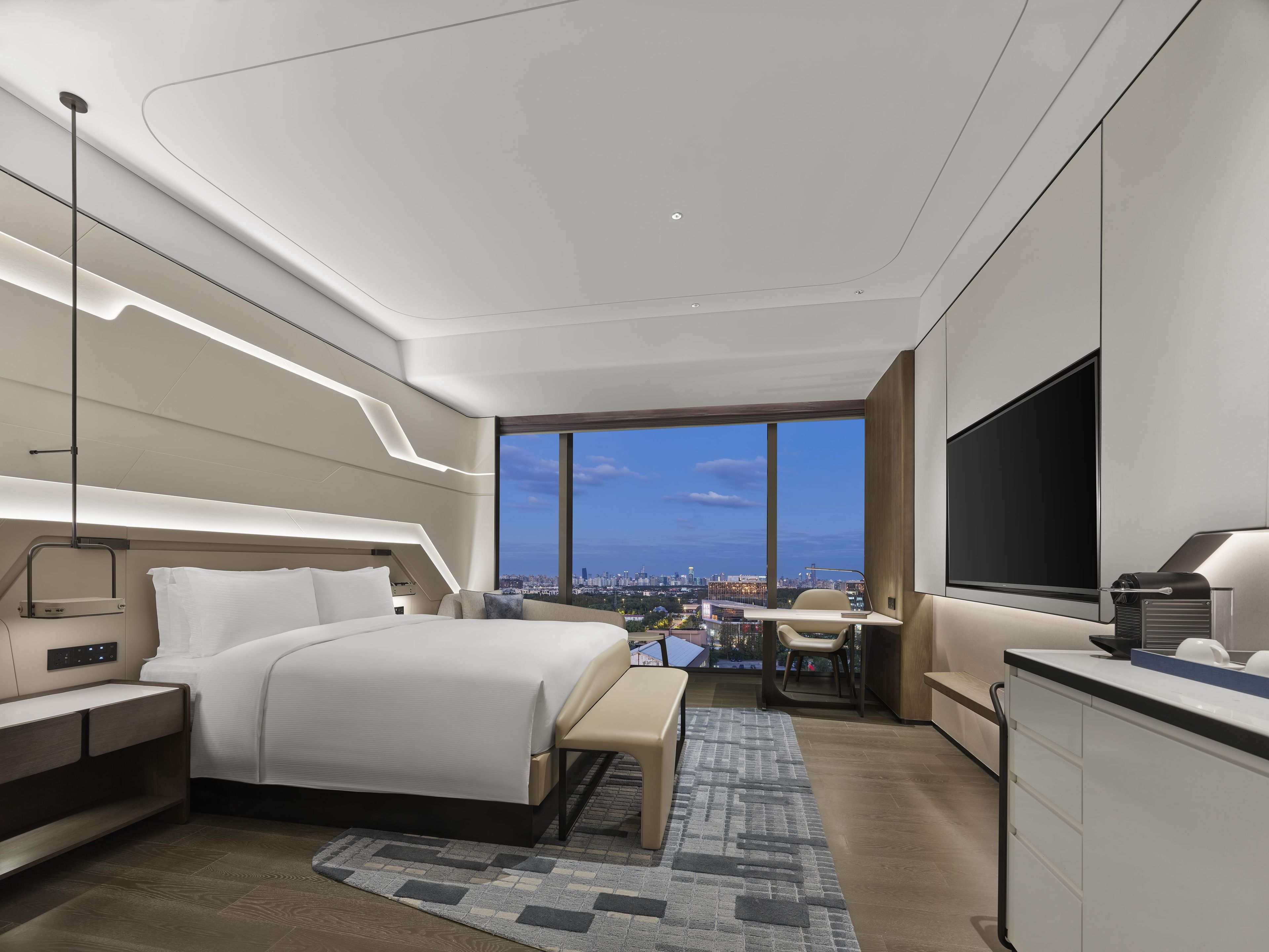 A modern hotel room with a large bed, bedside tables, a desk, a chair, and a TV.
