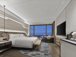 A modern hotel room with a large bed, bedside tables, a desk, a chair, and a TV.