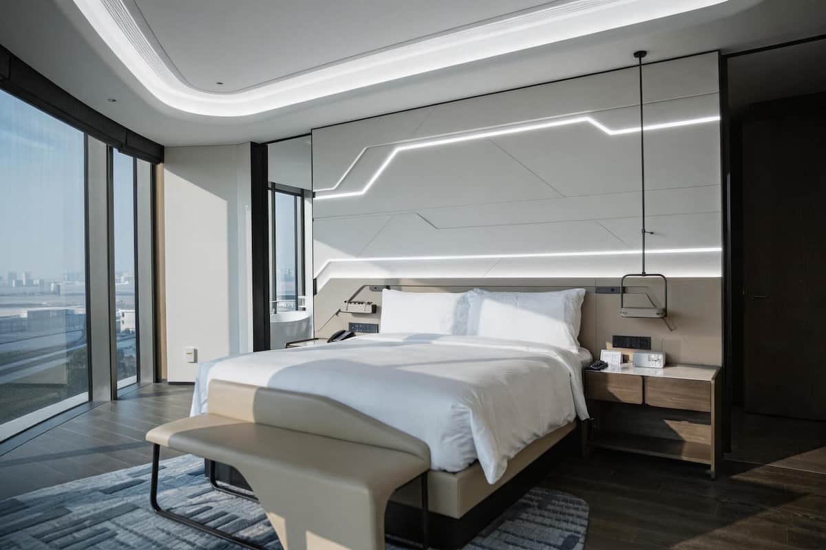 Habitación de hotel moderna con una cama grande, mesitas de noche, un banco, vistas a la ciudad y un diseño de techo único.
