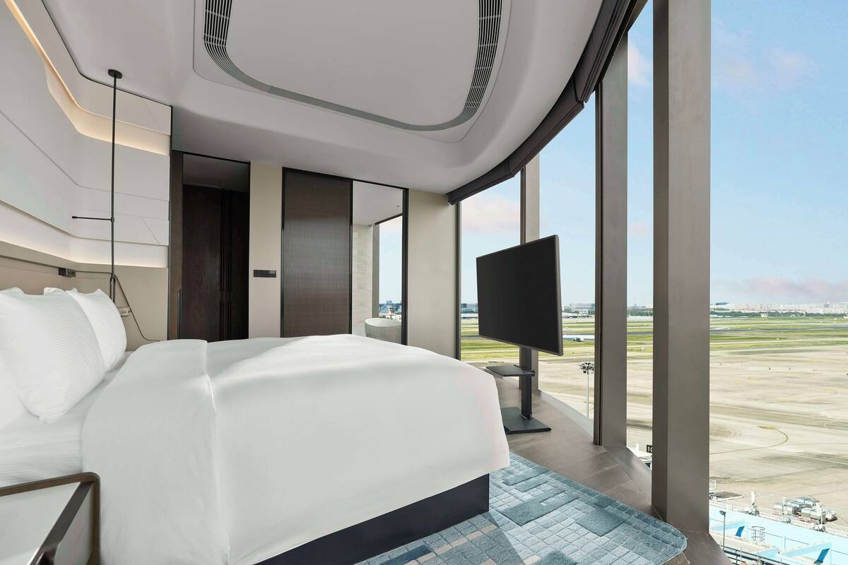 Ein modernes Hotelzimmer mit einem großen Bett, einem Flachbildfernseher und Blick auf einen Flughafen.