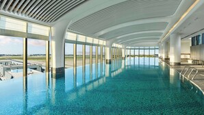 Una piscina interior amplia con diseño moderno, grandes ventanales que ofrecen vistas al aeropuerto y un techo con un original patrón de nervaduras.