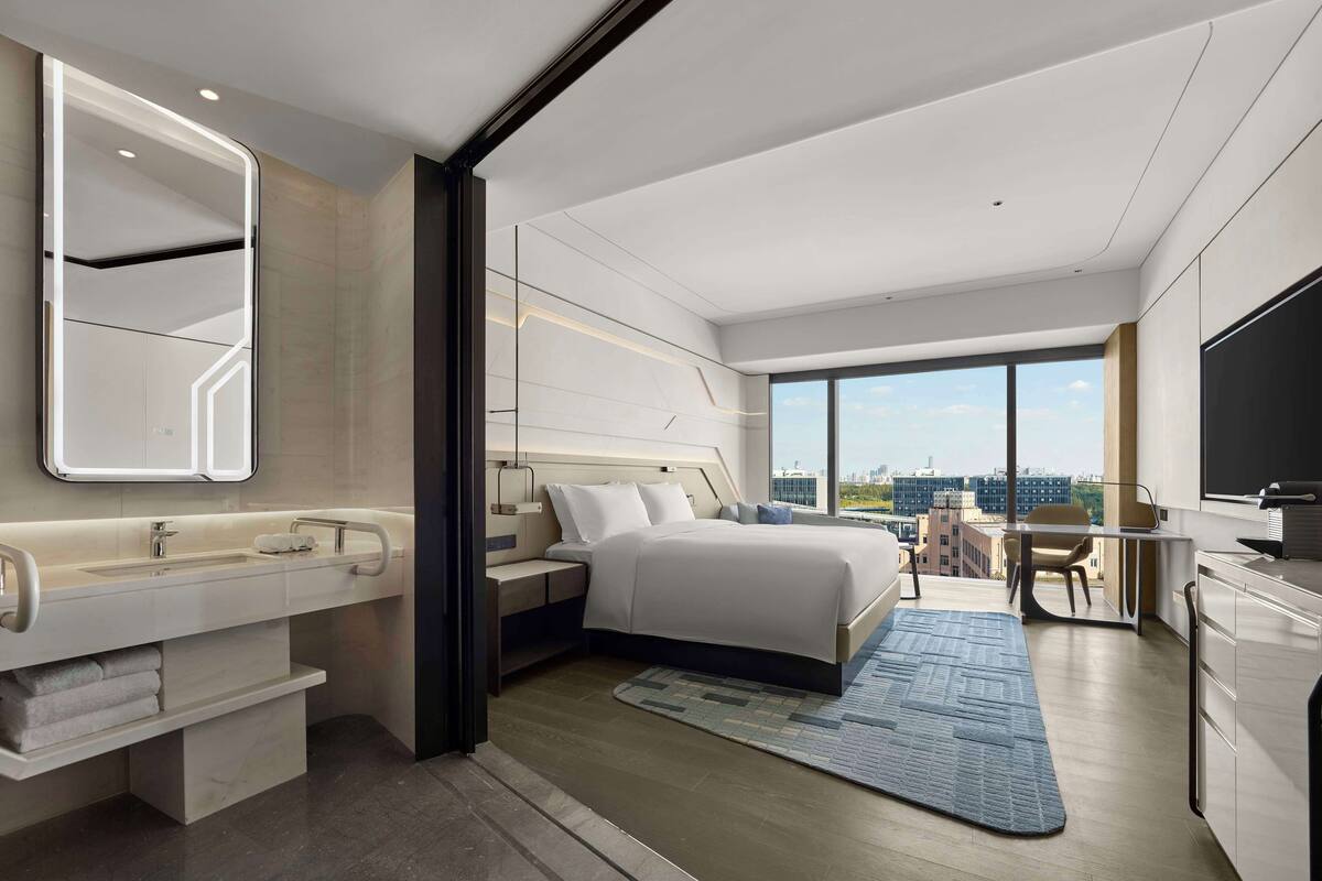 Ein modernes Hotelzimmer mit einem großen Bett, einem Schreibtisch, einem Sessel, einem Fernseher und einem Badezimmer mit Waschtisch und Spüle.