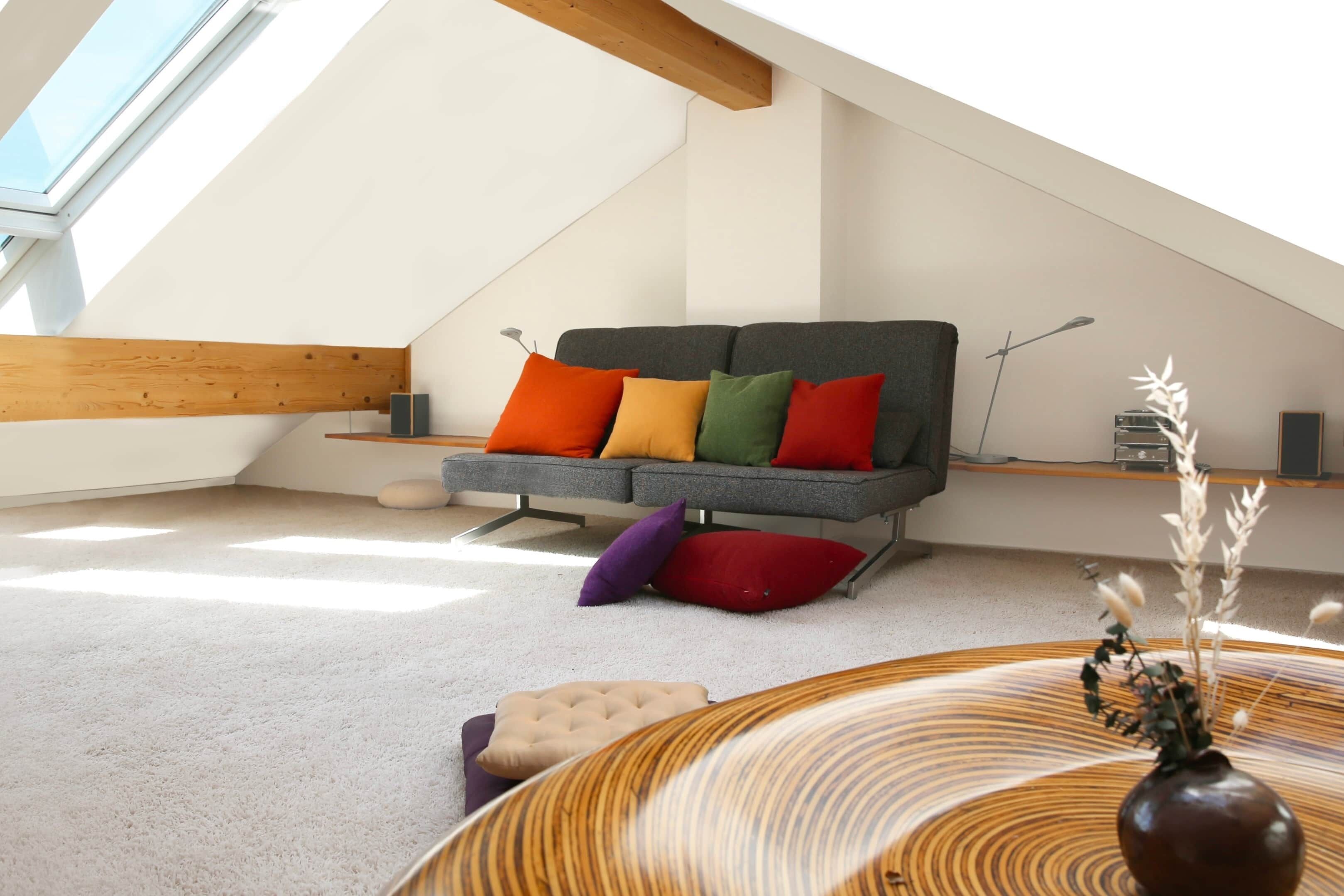 Un loft moderne avec un plafond en pente, un canapé gris agrémenté de coussins colorés, une table ronde en bois avec un vase, et une vue sur le ciel grâce à une lucarne.