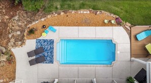 Una piscina rectangular con un borde de baldosas, un pequeño jardín con rocas y plantas, y una terraza de madera con sillas de descanso.