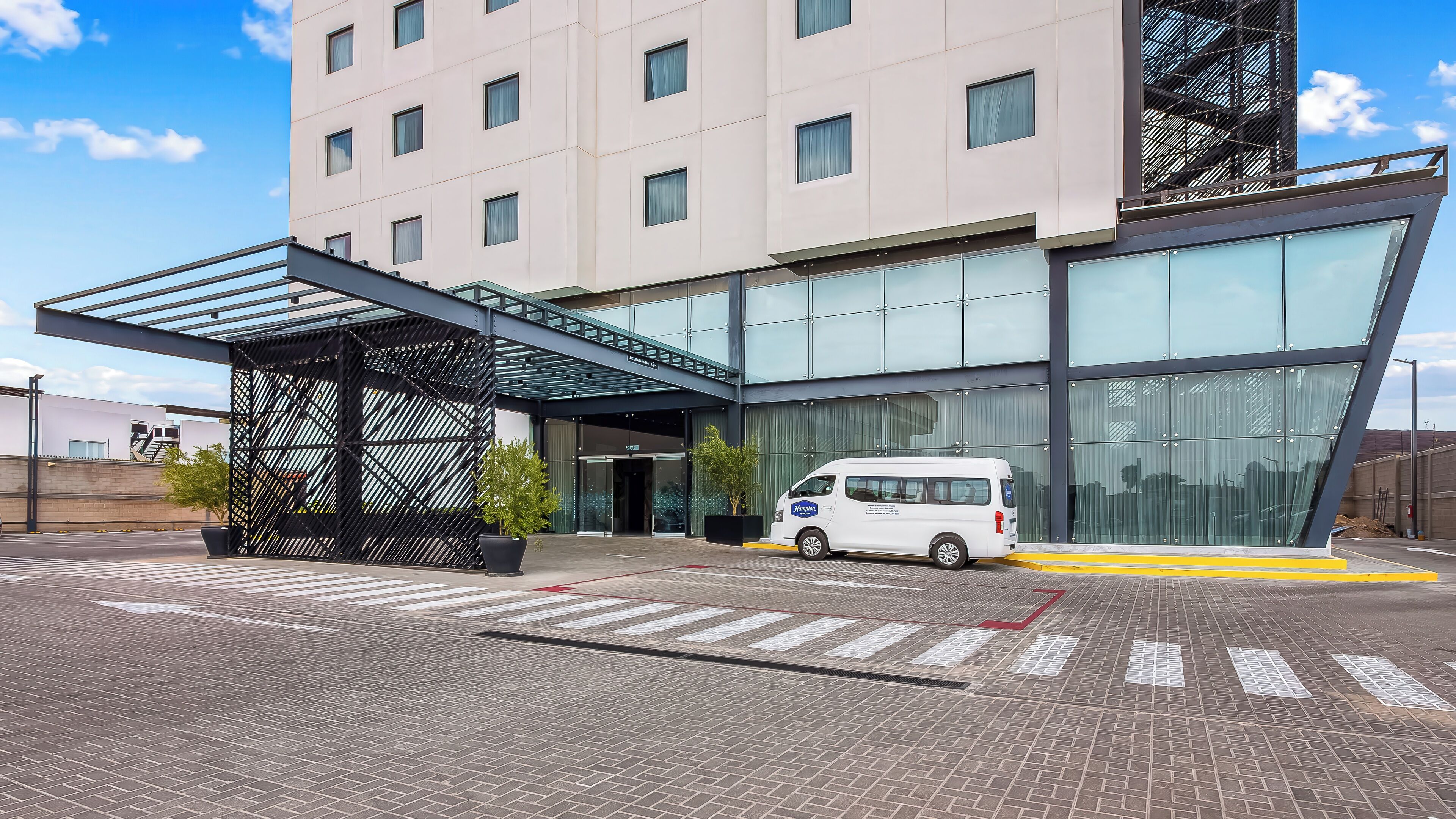 L’entrée moderne d’un hôtel, avec une allée couverte, un fourgon blanc garé devant et un ciel dégagé.