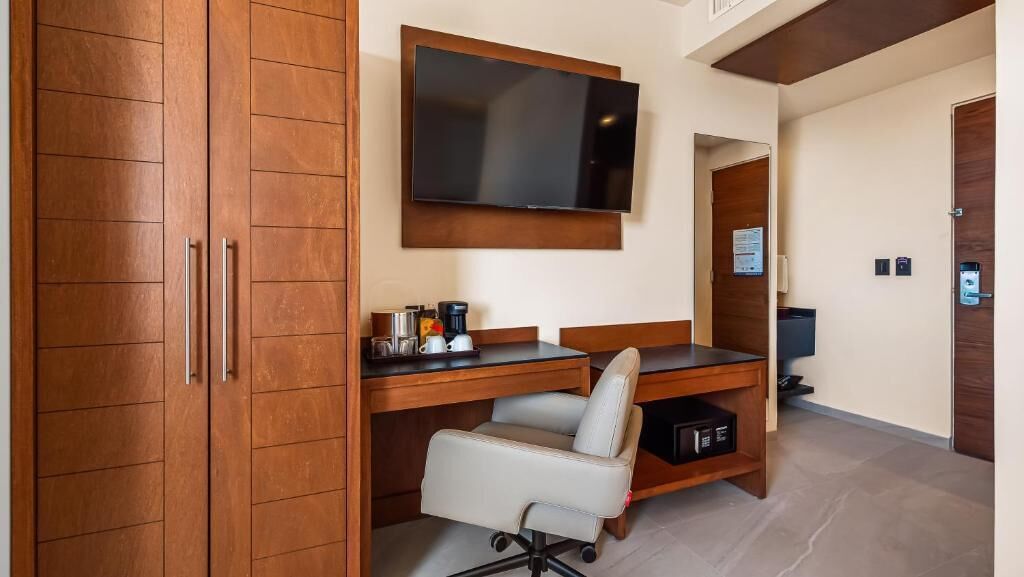 Quarto de hotel com armário de madeira, escrivaninha com cadeira, televisão de tela plana fixada acima da escrivaninha e prateleira pequena com cafeteira e outros objetos.
