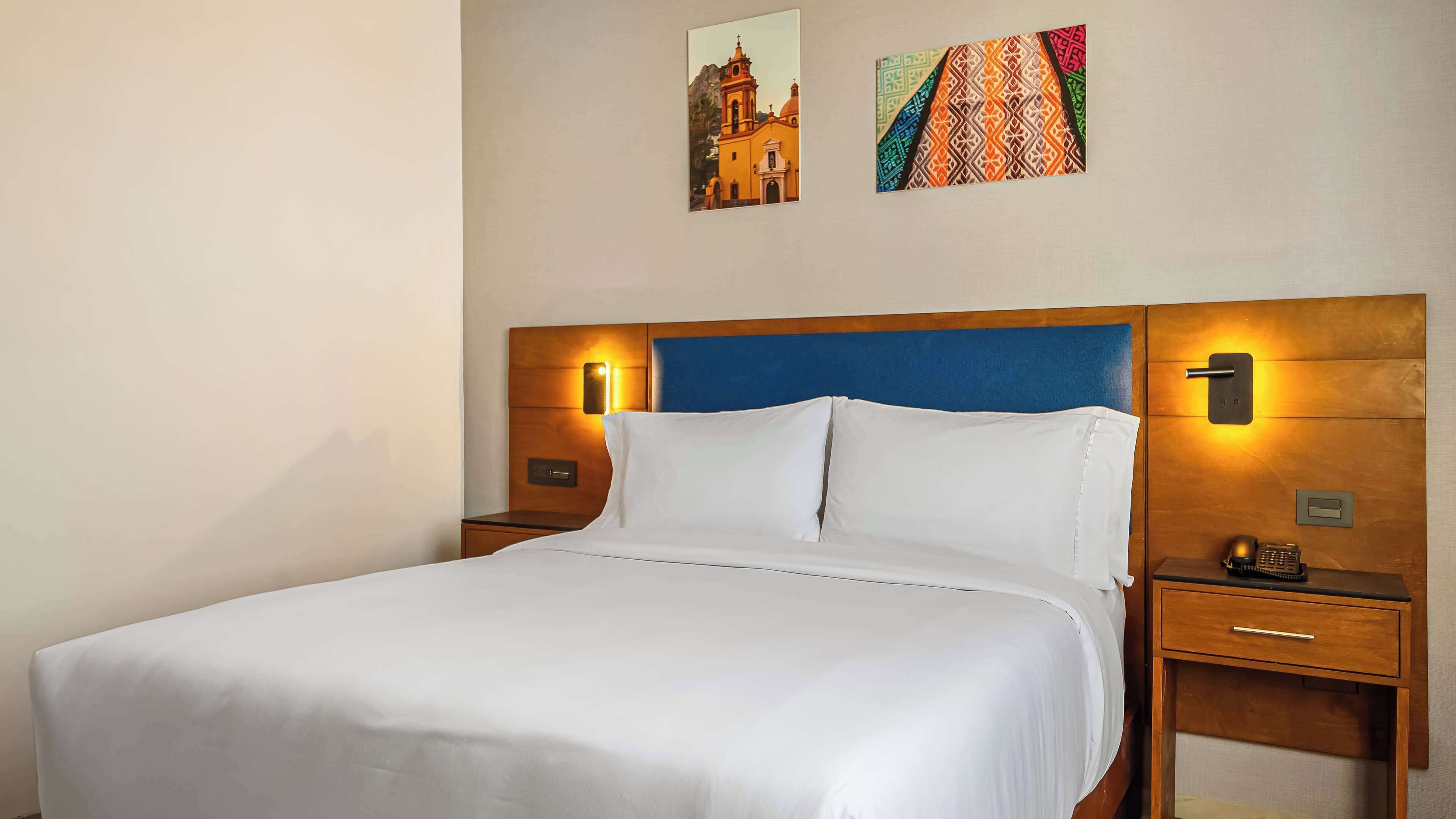 Quarto de hotel com uma cama grande, cabeceira de madeira, duas mesas de cabeceira e um criado-mudo.