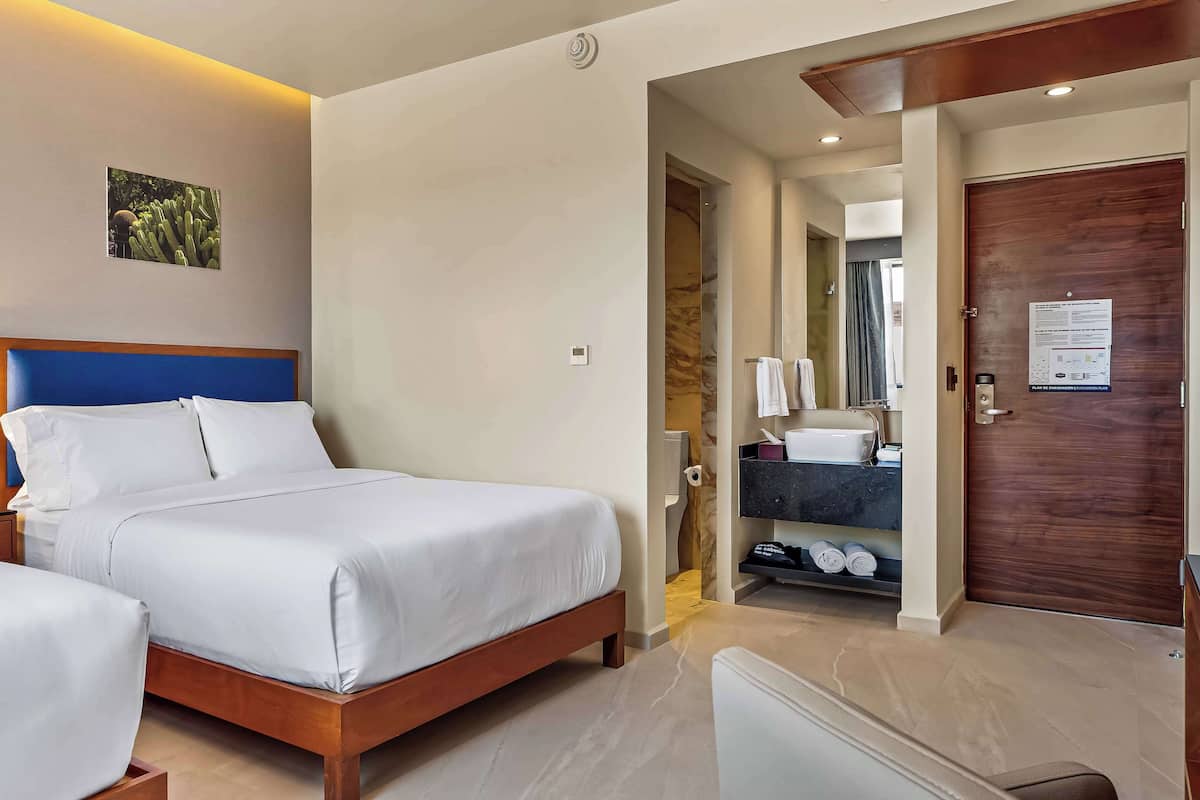 A hotel room with two beds, a wooden headboard, a nightstand, a bedside table, a wall-mounted light, a framed picture, and a bathroom visible through an open door.