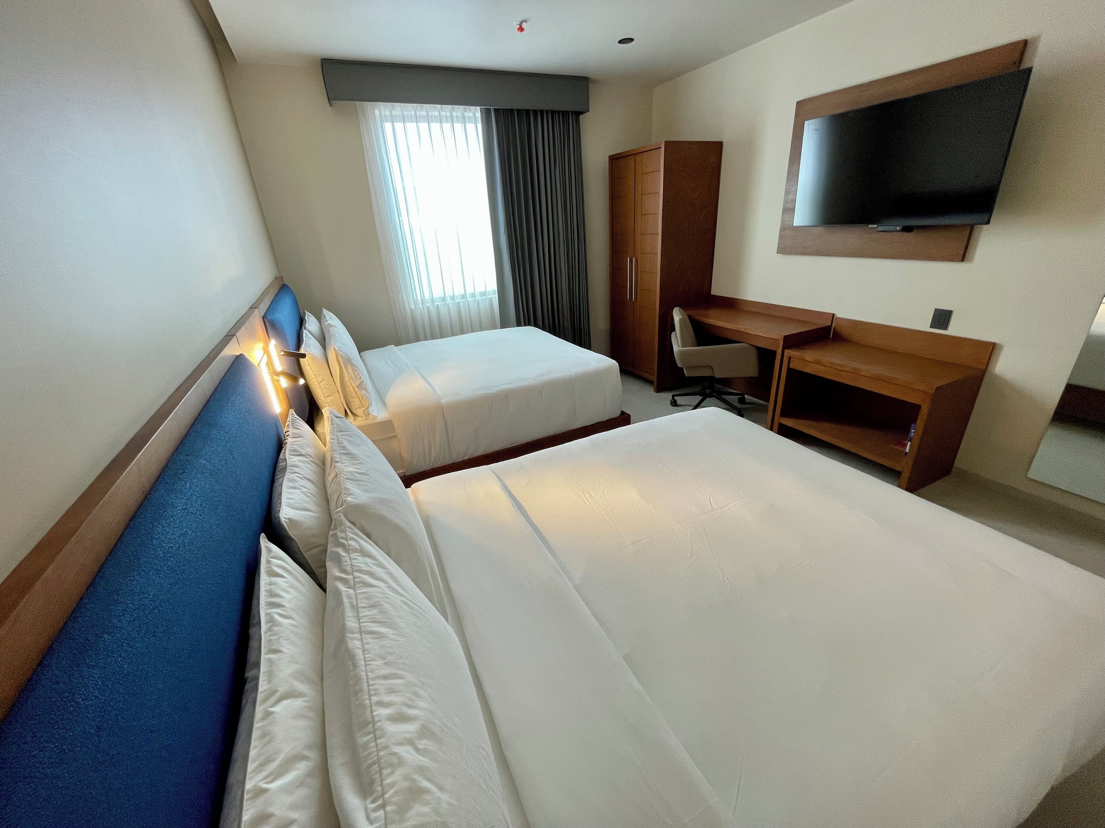 A hotel room with two beds, a desk, a chair, a TV, and a window with curtains.