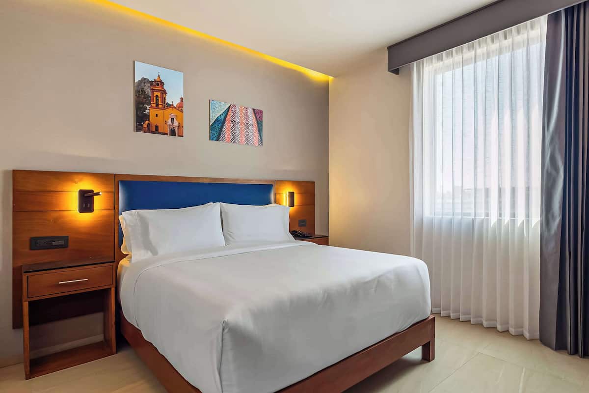 A hotel room with a large bed, a wooden nightstand, a bedside table, a wall-mounted lamp, and a window with sheer curtains.