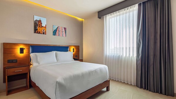 A hotel room with a large bed, a wooden nightstand, a bedside table, a wall-mounted lamp, and a window with sheer curtains. - Hampton By Hilton Queretaro Juriquilla (Querétaro)