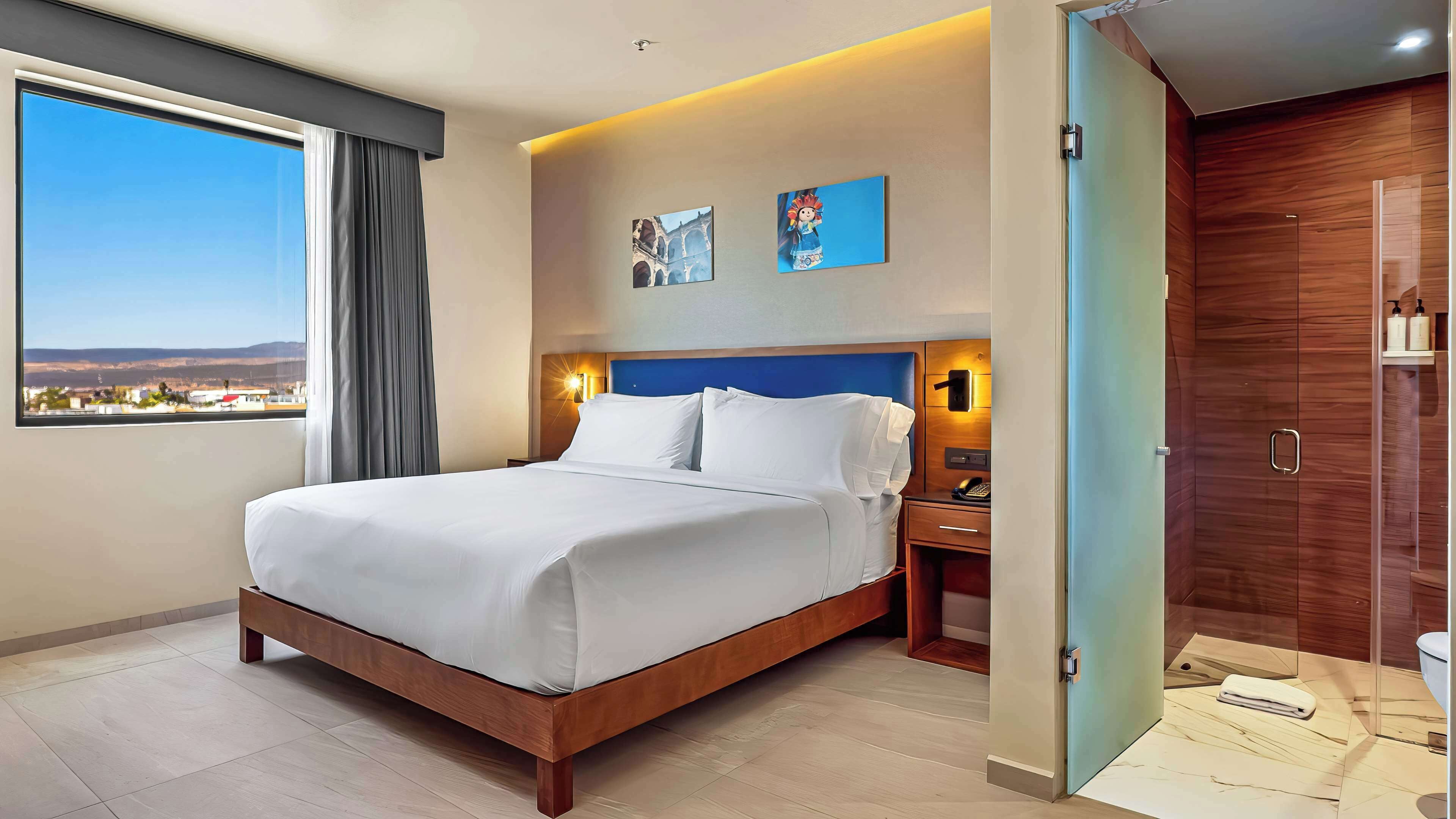 Quarto de hotel com uma cama grande, uma mesa de cabeceira, uma escrivaninha, banheiro com porta de madeira e vista para a cidade pela janela.