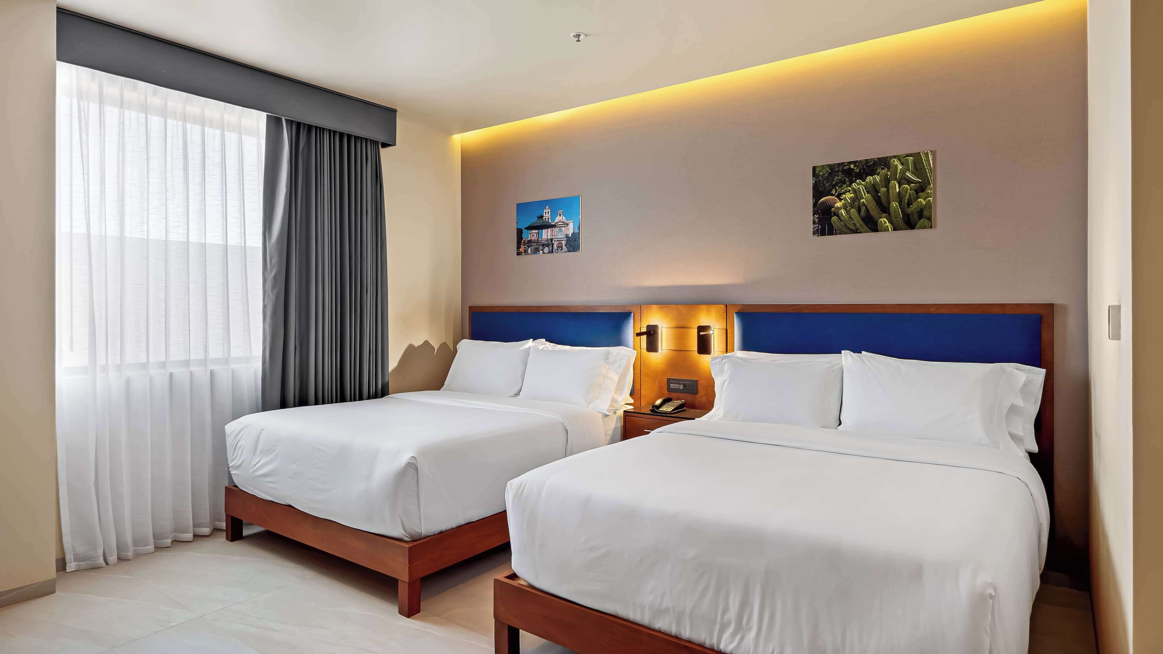 A hotel room with two beds, a large window with curtains, a wall-mounted lamp, and a painting.