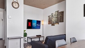 A modern living room with a flat-screen TV displaying streaming services, a sofa, a coffee table, and a dining table. - Loft 302 | Rooftop | Downtown (Rimouski)