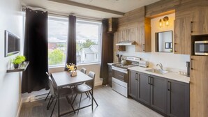 A modern kitchen with a dining area, wooden cabinets, stainless steel appliances, and a large window. - Loft 302 | Rooftop | Downtown (Rimouski)