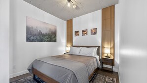 A modern bedroom with a large bed, a painting, and a series of wall-mounted lights. - Loft 302 | Rooftop | Downtown (Rimouski)