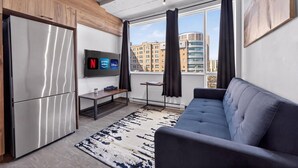 A modern living room with a sofa, a flat-screen TV, a refrigerator, and a window with curtains. - Loft 304 | Rooftop | Downtown (Rimouski)