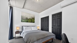 A bedroom with a bed, bedside lamps, a dark door, and a painting on the wall. - Loft 304 | Rooftop | Downtown (Rimouski)