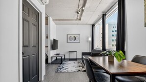 A modern room with a wooden dining table, black chairs, a TV on the wall, and a large window with curtains. - Loft 306 | Rooftop | Downtown (Rimouski)