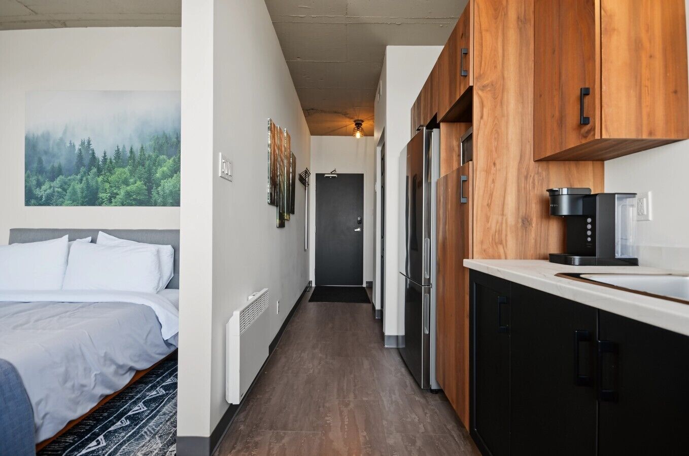 A hotel room with a bed, a painting, and a kitchenette with wooden cabinets and a coffee machine.