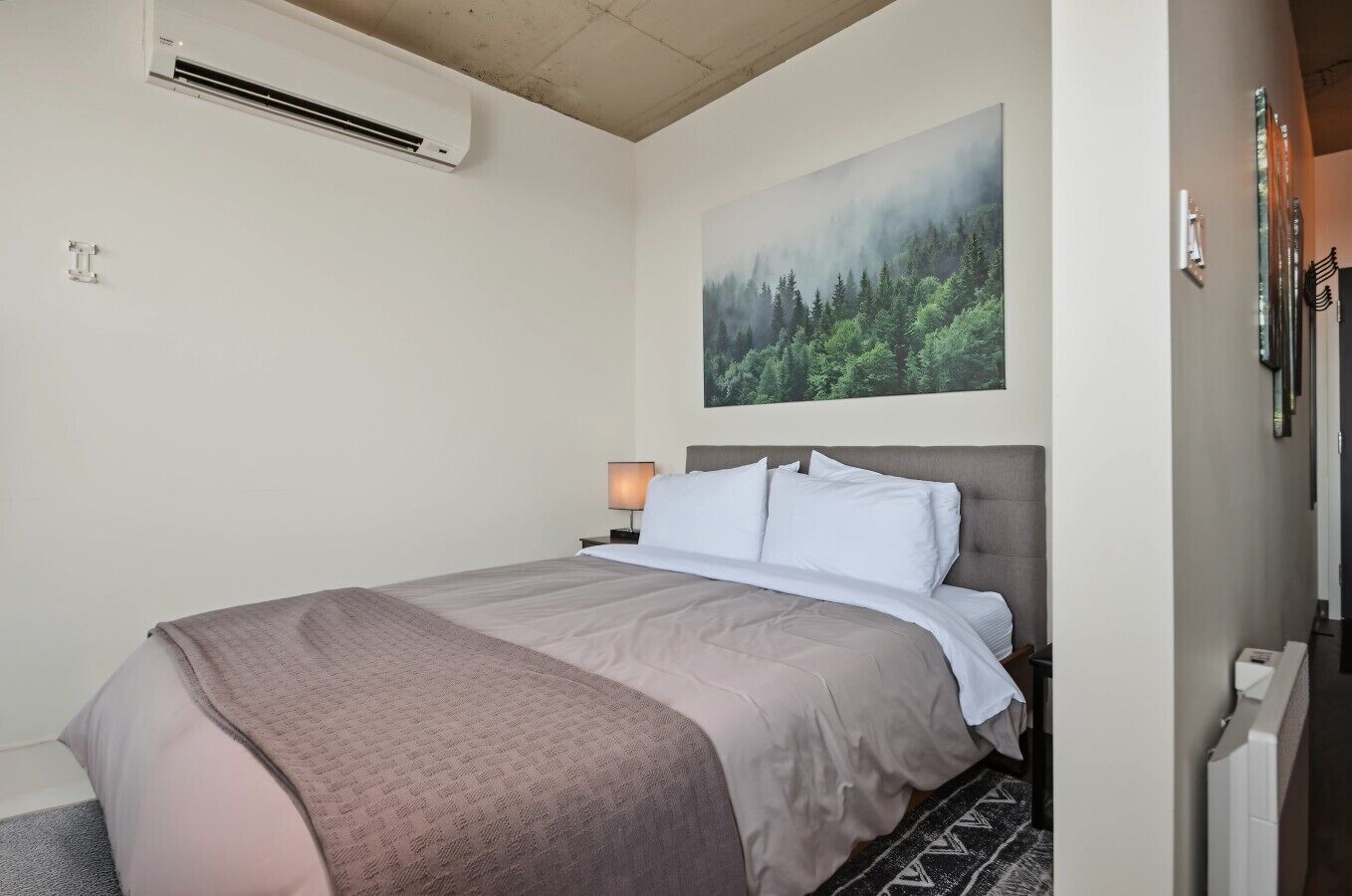 A hotel room with a bed, a nightstand, a lamp, an air conditioning unit, and a painting of a forest.