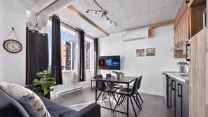 A modern living space with a sofa, dining table, chairs, a TV, and a kitchen area. - Loft 308 | Rooftop | Downtown (Rimouski)