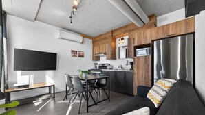 A modern living space with a flat-screen TV, dining table, chairs, a sofa, kitchen area with wooden cabinets, and a built-in microwave. - Loft 308 | Rooftop | Downtown (Rimouski)