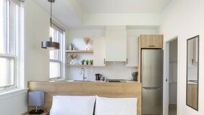 Un dormitorio moderno con cabecera de madera, cama con ropa de cama blanca, mesita de noche con lámpara y vistas a una cocina con refrigerador y armarios.