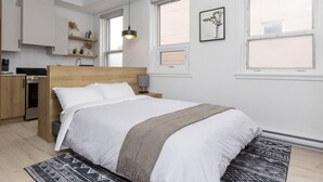 A modern bedroom with a large bed, a wooden headboard, a bedside table with a lamp, and a kitchen area in the background.