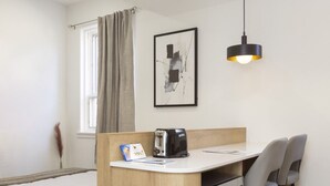 A compact room with a kitchenette, a bed, a window with curtains, a framed artwork, and a pendant light.