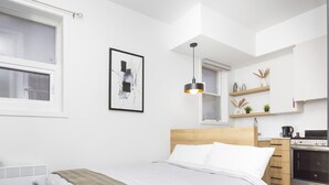 A modern bedroom with a wooden headboard, a bed with white linens, a black pendant light, a kitchen area with a stove, and a framed abstract painting on the wall.