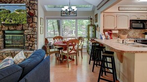 A spacious living room with a stone fireplace, a dining area with wooden chairs, and a kitchen with a large island and bar stools.