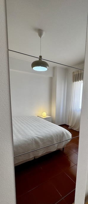 A bedroom with a bed, a bedside table with a lamp, a hanging light fixture, and a window with curtains.