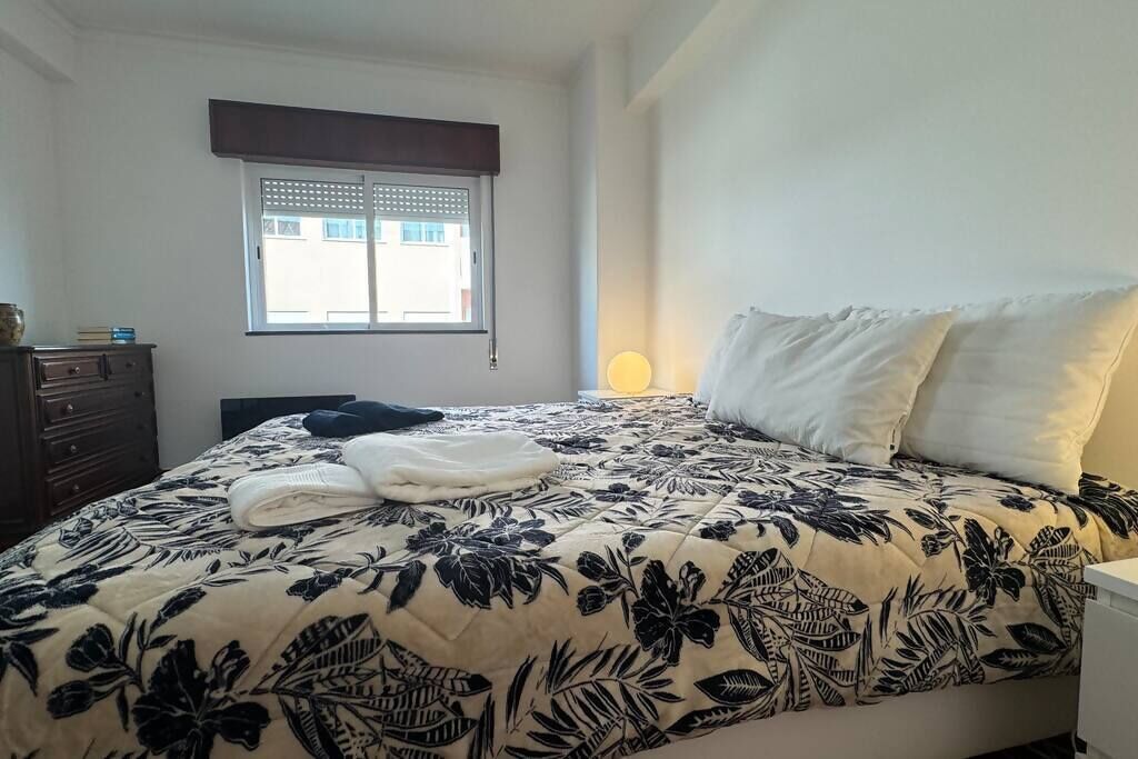 Een slaapkamer met een bed, een dressoir, een raam met jaloezieën en een bedlamp.