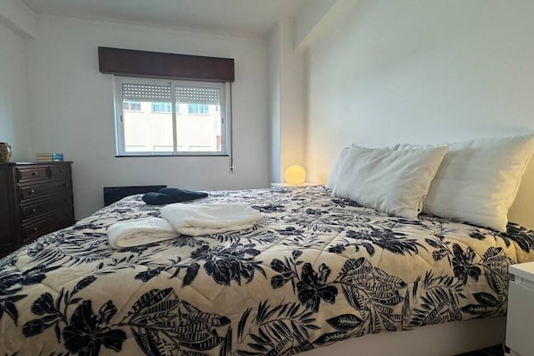 A bedroom with a bed, a dresser, a window with blinds, and a bedside lamp.