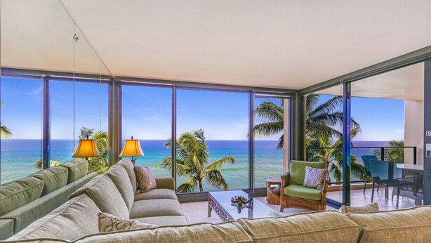 A modern living room with a large glass wall offering a view of the ocean. There are multiple seating areas, including a sofa and a chair, with a small table in the center. - Kaanapali Ocean Front 1 Bedroom! Mahana 617! (Lahaina)