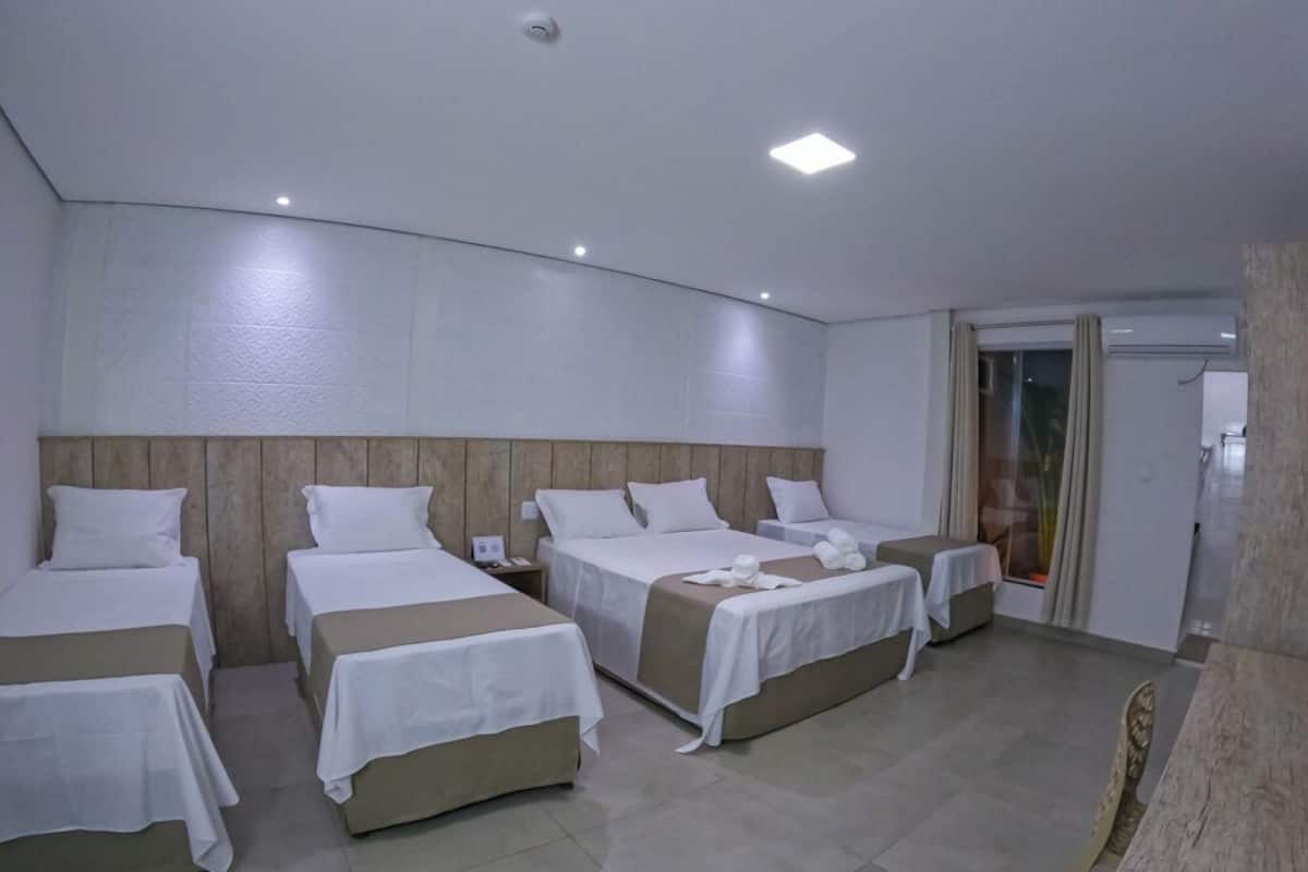 A hotel room with four beds arranged in a row, a large window with a view, and a door leading to a balcony.