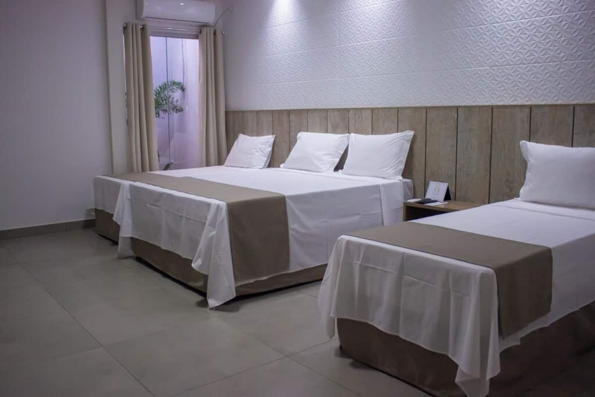 A hotel room with two beds, each with white linens and a beige bed runner. There is a window with curtains, a wooden headboard, and a small table with a phone and a notepad.