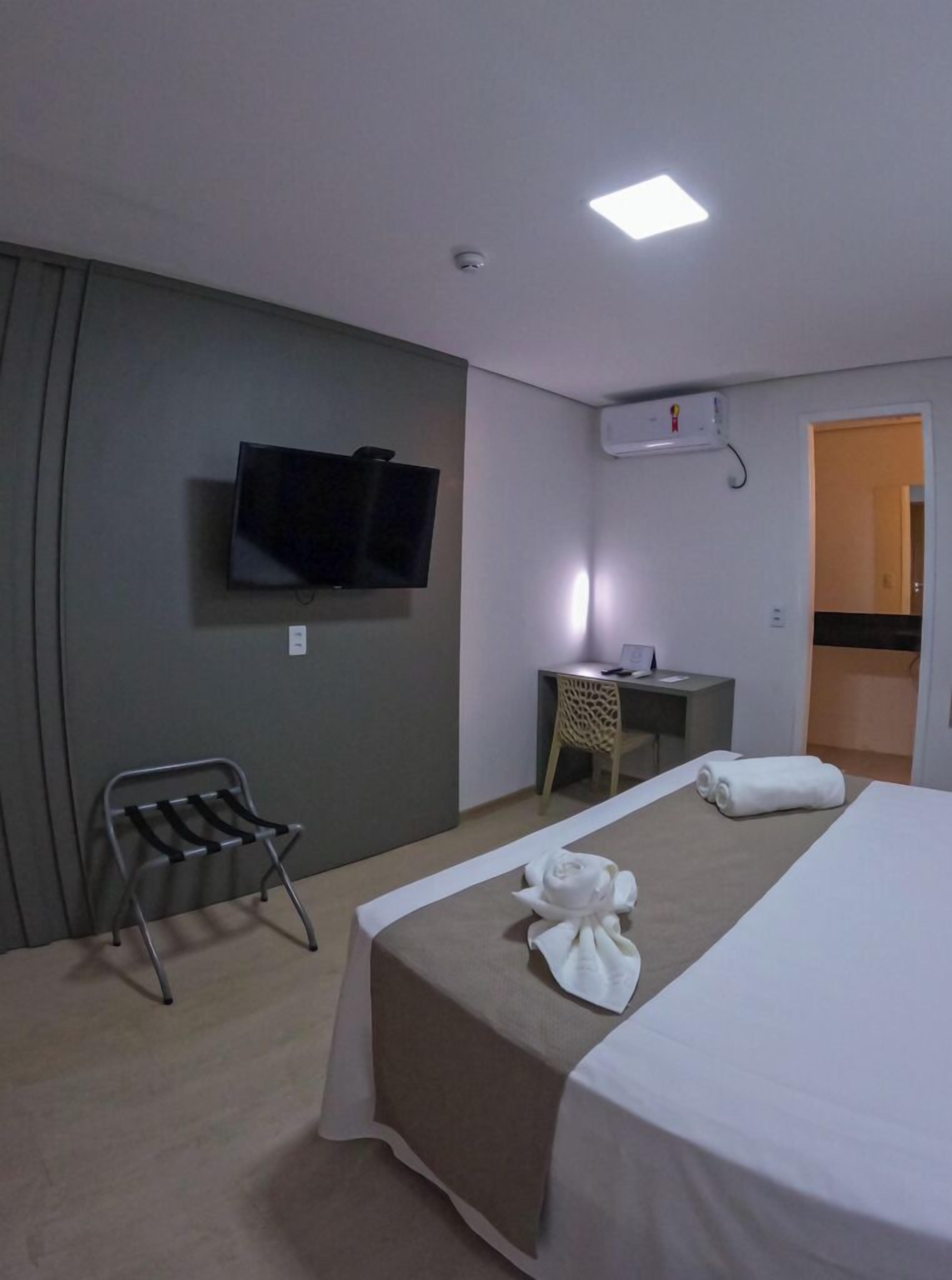 A hotel room with a bed, a desk, a chair, a TV mounted on the wall, and an air conditioning unit.