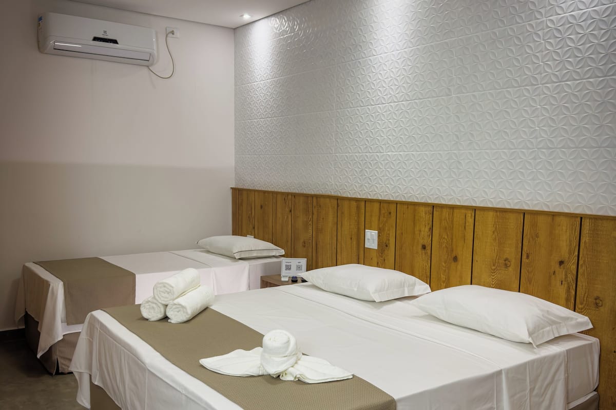 A hotel room with two beds, each with white linens and a brown bed runner. There is a wooden headboard, a wall-mounted air conditioning unit, and a small bedside table with a lamp.